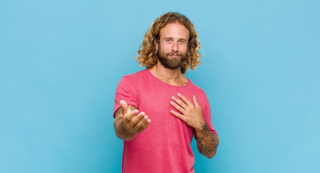 Blonde man feeling happy and in love, smiling with one hand next to heart and the other stretched up front