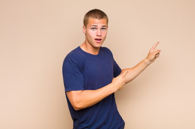 Blonde  man feeling shocked and surprised, pointing to copy space on the side with amazed, open-mouthed look