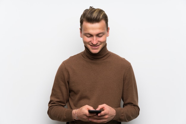 Blonde man over isolated white wall sending a message with the mobile