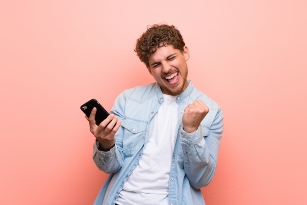 Blonde man over pink wall celebrating a victory with a mobile