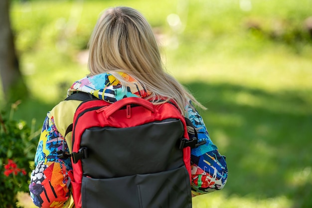 Blonde middleaged woman in bright clothes with a backpack in the park on a sunny day rear view