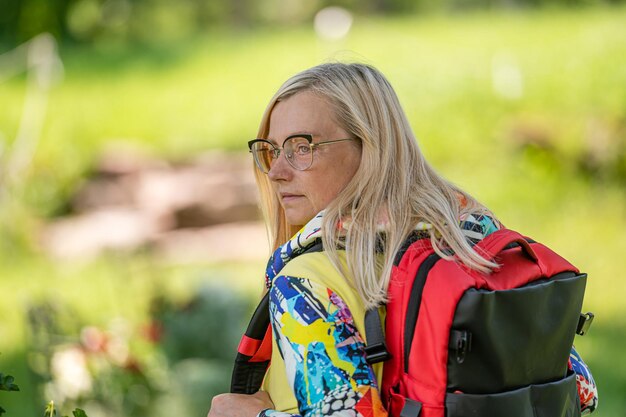 Blonde middleaged woman in bright clothes with a backpack in the park on a sunny day rear view