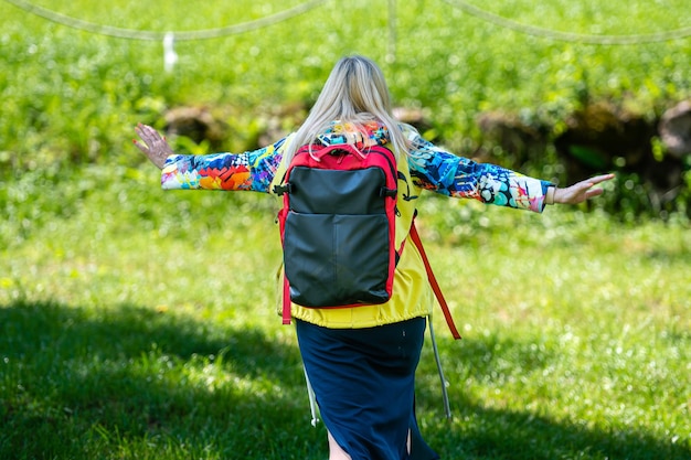 Blonde middleaged woman in bright clothes with a backpack in the park on a sunny day rear view