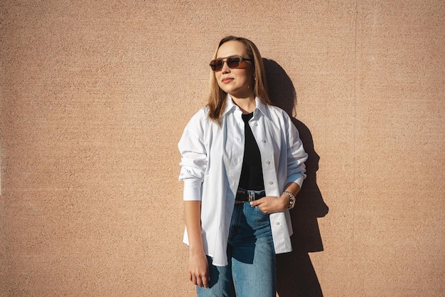 The blonde model wearing sunglasses stands against the beige wall