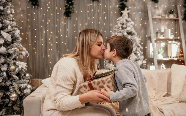 Blonde mom in a knitted suit is sitting on the couch receiving a gift from her son on a New Year's background