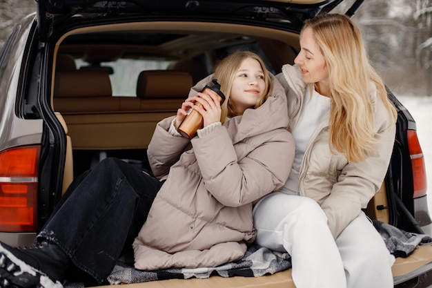 Blonde mother and teen daughter sitting in open car's trunk in winter forest