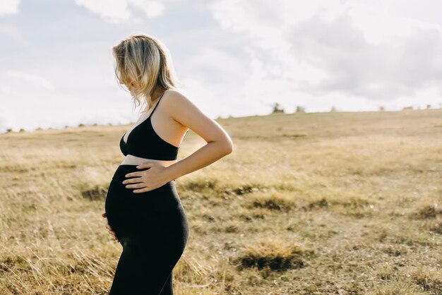 Blonde pregnant woman wearing all black standing in an open field holding hands on belly