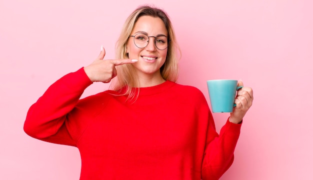 Photo blonde pretty woman smiling confidently pointing to own broad smile coffee cup concept