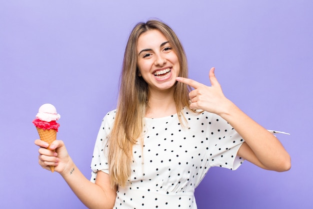 Blonde pretty woman smiling confidently pointing to own broad smile, positive, relaxed, satisfied attitude