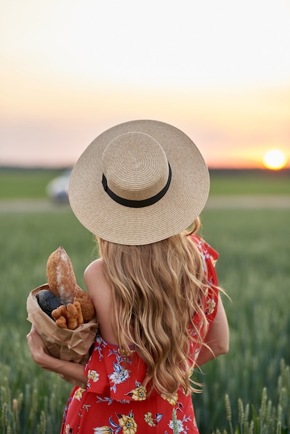 A blonde in a red dress and a straw hat with French bread in her hands on a wheat field at sunset