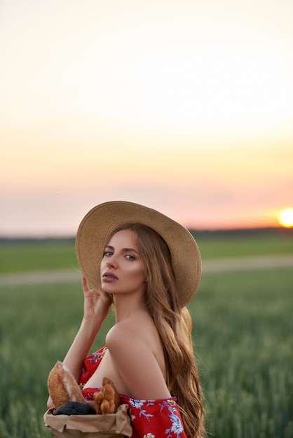 A blonde in a red dress and a straw hat with French bread in her hands on a wheat field at sunset