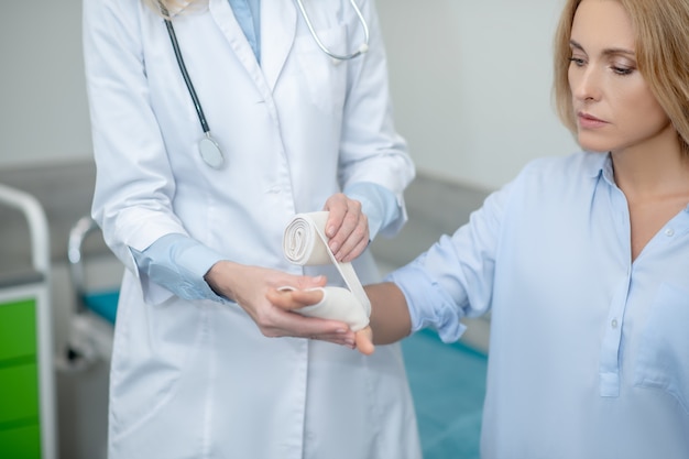 Blonde sad woman sitting with outstretched arm and doctor hands applying elastic bandage