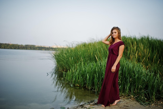 Blonde sensual barefoot woman in red marsala dress posing against lake with reeds