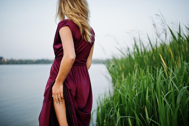 Blonde sensual woman in red marsala dress standing in water of lake with reeds and show her nude leg