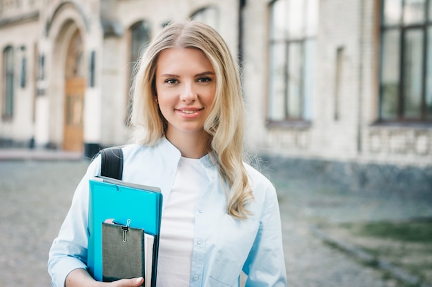 blonde student girl is smiling and holding a folder and a notebook in her hands in university