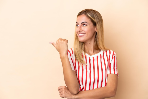 Blonde Uruguayan girl isolated on beige background pointing to the side to present a product