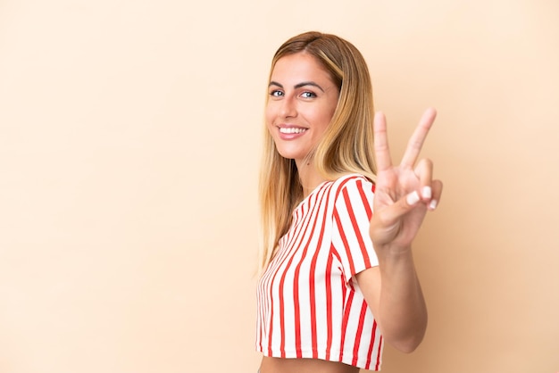 Blonde Uruguayan girl isolated on beige background smiling and showing victory sign