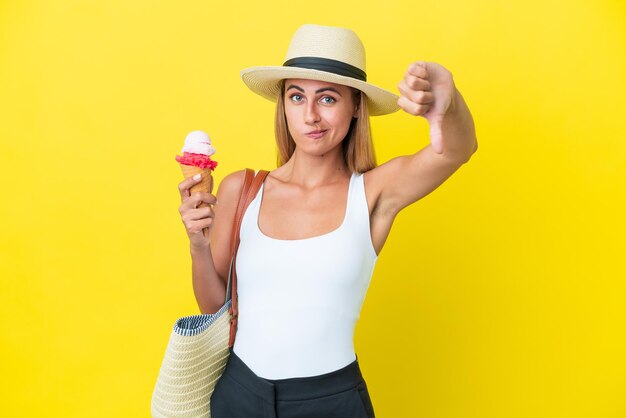 Blonde Uruguayan girl in summertime holding ice cream isolated on yellow background showing thumb down with negative expression