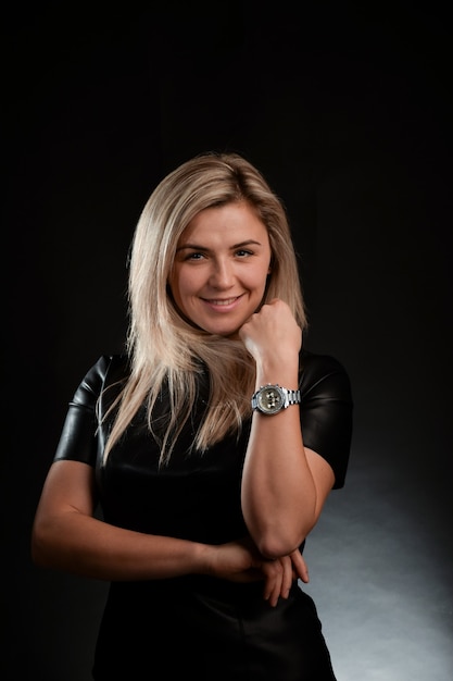 A blonde white race girl with long hair smiles and looks into the camera lens. Dressed in a black dress. The background is dark.