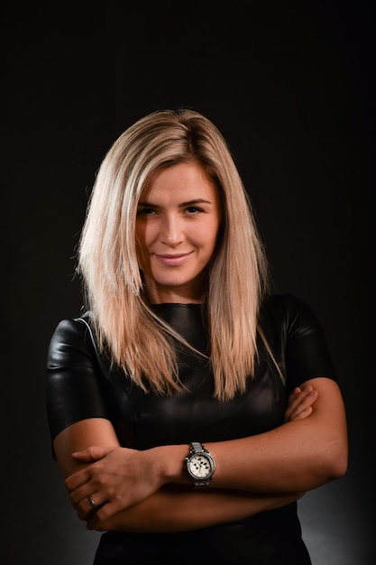 A blonde white race girl with long hair smiles and looks into the camera lens. Dressed in a black dress. The background is dark.