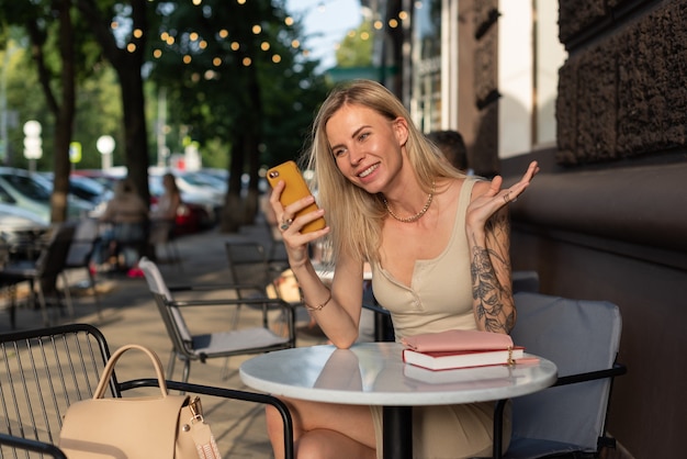 A blonde with a tattoo on her arm sits in a summer cafe and emotionally talks on the phone