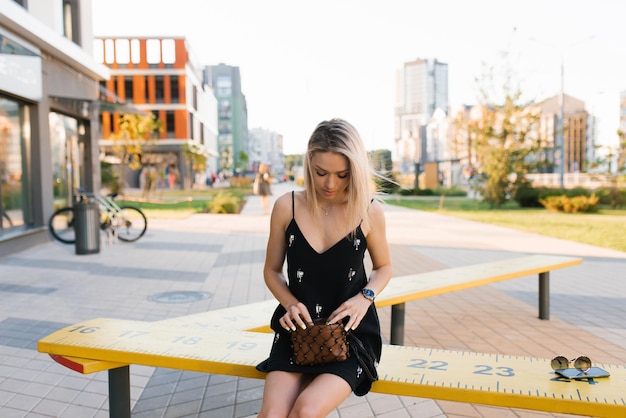 Blonde woman in a black dress is sitting on a bench in the city and is looking for something