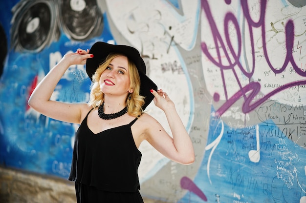 Blonde woman on black dress, necklaces and hat against graffiti wall.