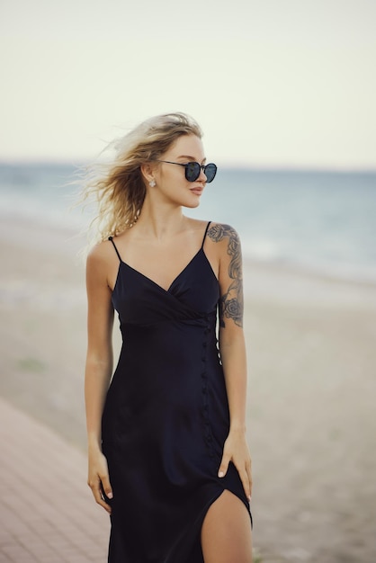 Blonde woman in black dress outdoors against seashore on the background during summer beach