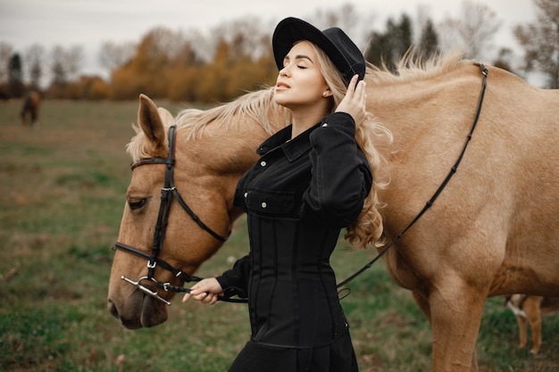 Blonde woman and brown horse standing in the field. Woman wearing black clothes and hat. Woman touching the horse.