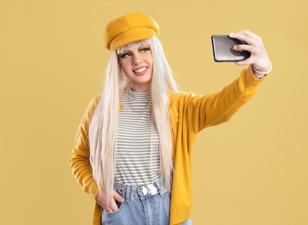 Blonde woman in cap and yellow jacket making selfie with phone on yellow backgroundBlonde woman in cap and yellow jacket taking a photo with the smartphone on yellow background