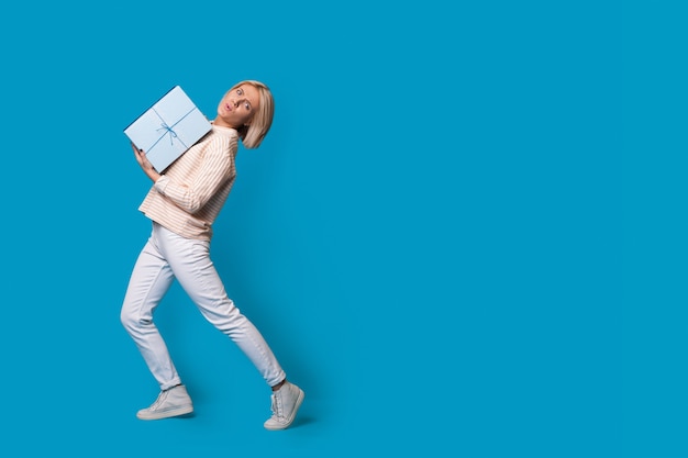 Blonde woman carrying a box with gift on a blue wall studio advertising something