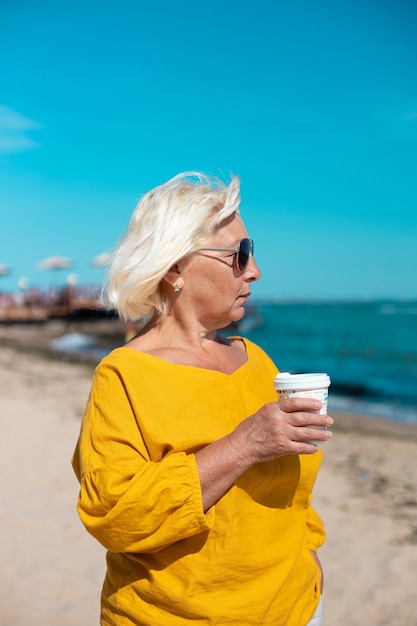 Blonde woman in cotton blouse holding paper cup with coffee drink while walking on the beach