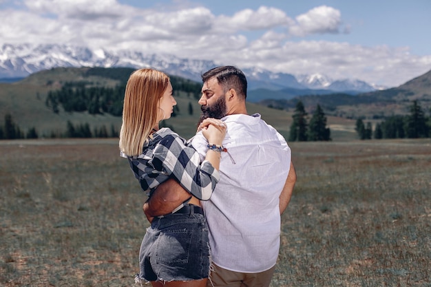 Blonde woman and a dark-haired bearded man are standing in an embrace and looking at each other against backdrop of a picturesque forest landscape with mountains, trees and a sky with several clouds.