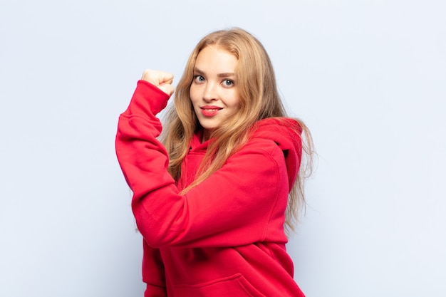 Blonde woman feeling happy, satisfied and powerful, flexing fit and muscular biceps, looking strong after the gym