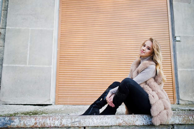 Blonde woman at fur coat against wall with shutters.