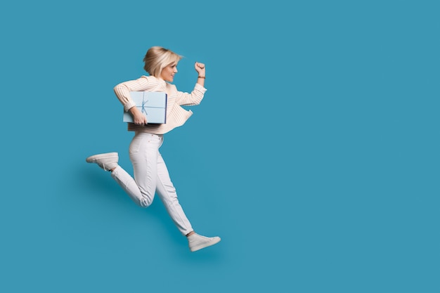 Blonde woman is running on a  wall with blue free space holding a present box in hands