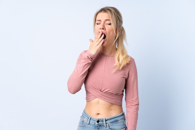 Blonde woman over isolated blue yawning and covering wide open mouth with hand