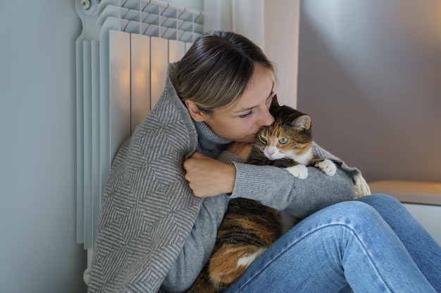 Blonde woman leaning on room heating battery kisses domestic cat sitting together on floor