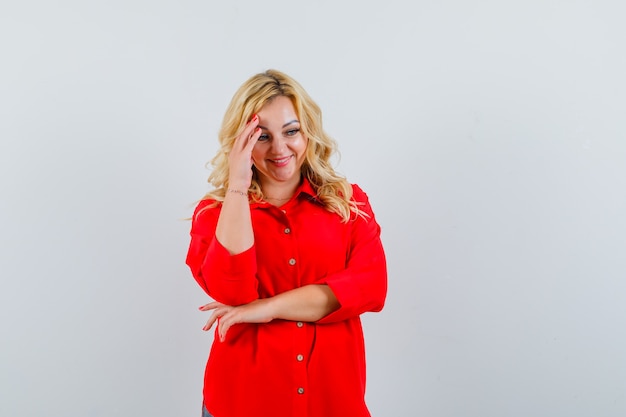 Blonde woman in red blouse putting hand on head, thinking about something and looking pretty , front view.