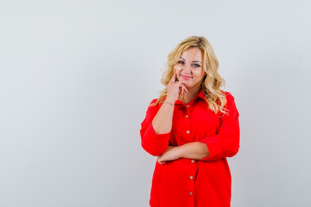 Blonde woman in red blouse putting index finger on cheek and looking pretty , front view.