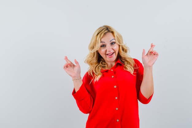 Blonde woman in red blouse standing fingers crossed and looking happy , front view.