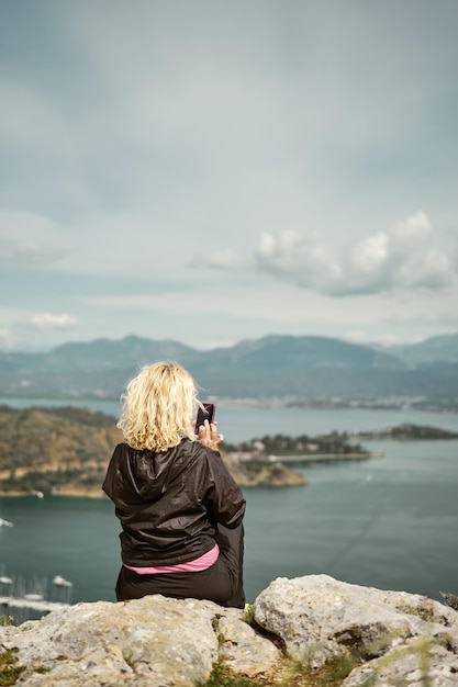 Blonde woman sits on a high rock overlooking the marina and the bay in the Aegean Sea on a cloudy day and shoots a video on a smartphone Vertical frame travel and vacation concept