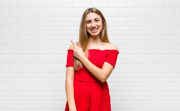 Blonde woman smiling cheerfully, feeling happy and pointing to the side and upwards, showing object in copy space