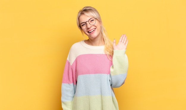 Photo blonde woman smiling happily and cheerfully, waving hand, welcoming and greeting you, or saying goodbye