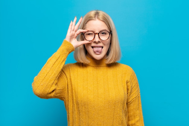 Blonde woman smiling happily with funny face, joking and looking through peephole, spying on secrets