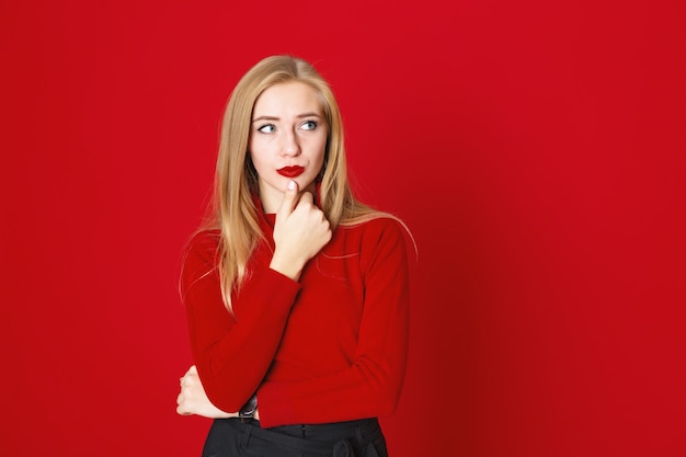 Blonde woman standing on a red background wearing casual  sweater holding herself by chin