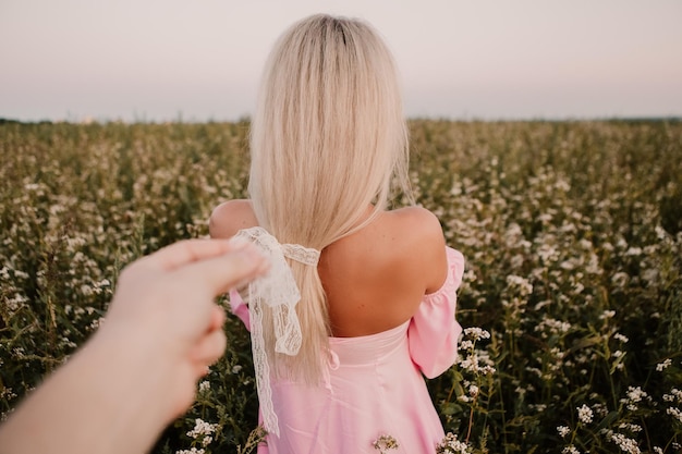 Blonde woman walking in the big endless field of daisies in summer evening. Lady wear pink dress