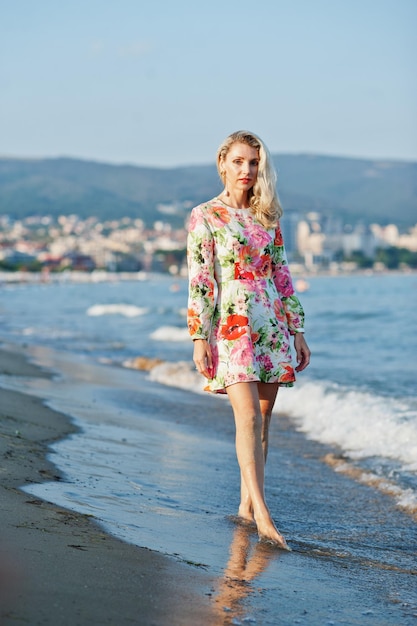 Blonde woman wearing dress standing on sand beach at sea shore enjoying view of sunset