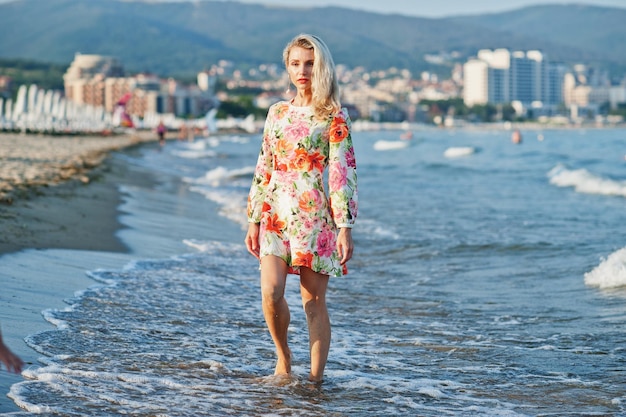 Blonde woman wearing dress standing on sand beach at sea shore enjoying view of sunset