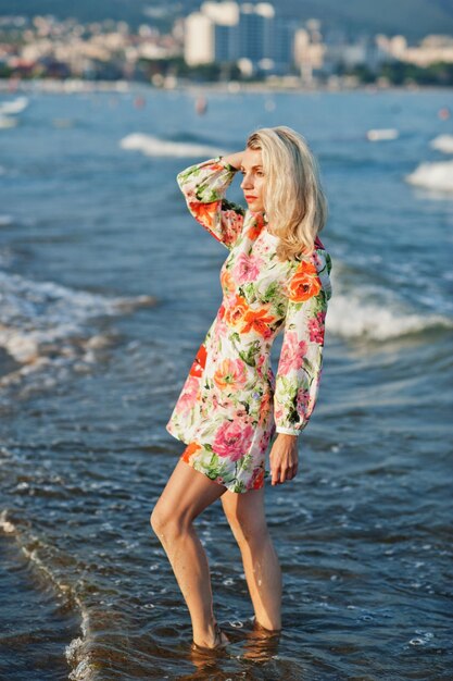 Blonde woman wearing dress standing on sand beach at sea shore enjoying view of sunset